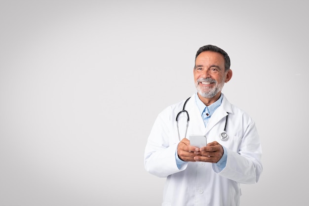 Médecin senior européen souriant en blouse blanche avec stéthoscope discutant au téléphone en regardant vide