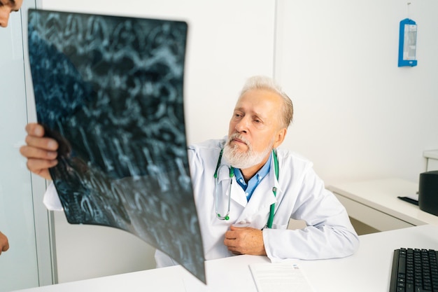 Médecin de sexe masculin adulte adulte pensif regardant l'image de la colonne vertébrale IRM du patient assis au bureau avec un ordinateur dans la salle du cabinet médical, pensant au diagnostic, exprimant son inquiétude face à la maladie.