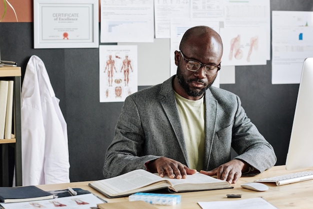 Médecin de sexe masculin africain examinant les maladies dans le livre assis à table pendant le travail au bureau