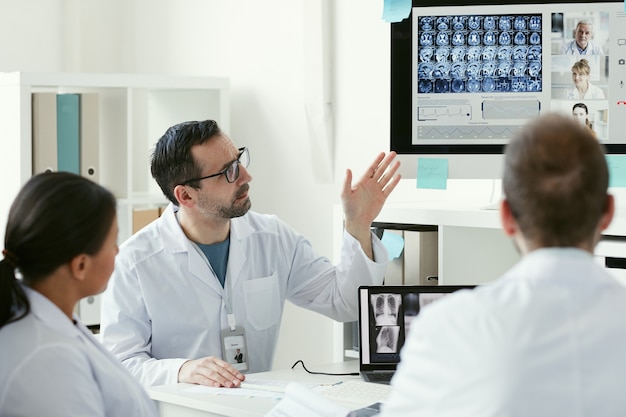 Médecin de sexe masculin d'âge mûr pointant sur des images radiographiques sur écran d'ordinateur et en discuter avec ses collègues au bureau