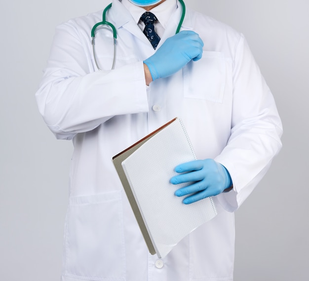 Médecin de sexe masculin en blouse blanche avec des boutons, portant des gants stériles bleus, tenant un cahier en papier