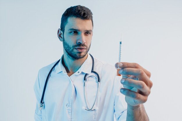 Médecin de sexe masculin européen concentré regardant la seringue. Jeune homme barbu avec stéthoscope portant une blouse blanche. Isolé sur fond turquoise. Prise de vue en studio