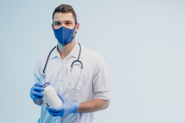 Médecin de sexe masculin européen tenant une bouteille de pilules. Jeune homme avec stéthoscope portant une blouse blanche, un masque de protection et des gants en latex. Isolé sur fond gris. Tournage en studio. Espace de copie