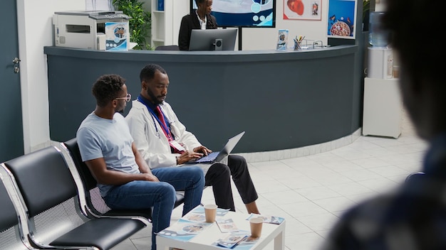 Médecin de sexe masculin expliquant le diagnostic de la maladie au patient dans le hall d'accueil de l'hôpital, utilisant un ordinateur portable pour montrer les résultats des soins de santé et donner un traitement médical. Médecin et personne assise dans la salle d'attente.