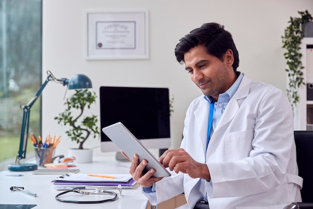 Médecin de sexe masculin ou généraliste portant un manteau blanc assis au bureau au bureau à l'aide d'une tablette numérique