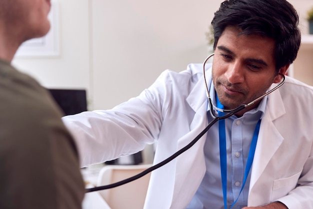 Médecin de sexe masculin ou généraliste portant un manteau blanc examinant un jeune homme écoutant la poitrine avec un stéthoscope
