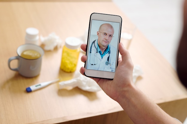 Médecin de sexe masculin mature sur écran de smartphone regardant le patient et donnant des recommandations médicales à un jeune homme malade décrivant ses symptômes