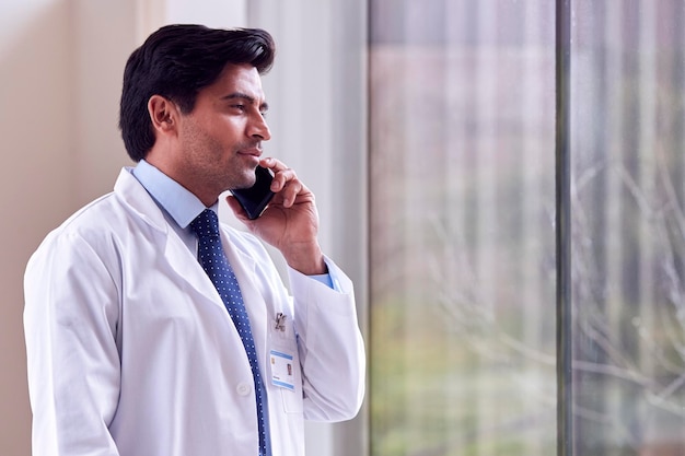 Médecin de sexe masculin portant un manteau blanc debout dans le couloir de l'hôpital parlant sur téléphone mobile