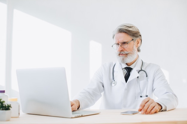 Médecin de sexe masculin senior pense et vérifie ses dossiers médicaux des patients sur un ordinateur portable dans la salle médicale