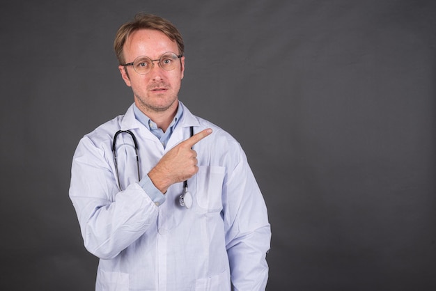 Médecin de sexe masculin souriant avec stéthoscope en manteau médical pointant le doigt sur fond gris