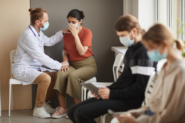 Médecin de sexe masculin soutenant le patient pendant qu'elle pleure, ils sont assis dans le couloir de l'hôpital avec d'autres personnes