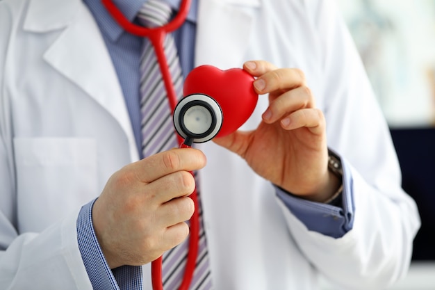 Médecin de sexe masculin tenant un coeur rouge et mettant la tête du stéthoscope près d'elle gros plan. Aide médicale cardiologie soins santé prophylaxie prévention assurance chirurgie et réanimation concept