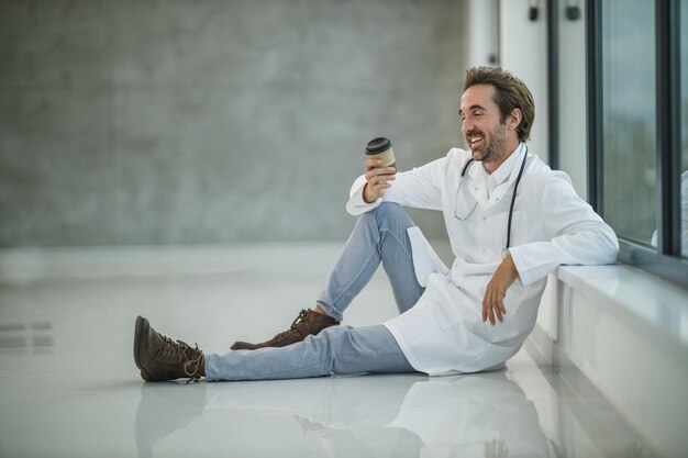 Un médecin souriant assis sur le sol près d'une fenêtre tout en prenant une pause-café rapide dans un couloir vide de l'hôpital pendant la pandémie de Covid-19.