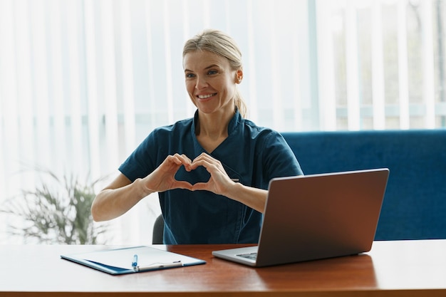 Médecin souriant travaillant sur un ordinateur portable et montrant le cœur avec les mains dans un cabinet médical