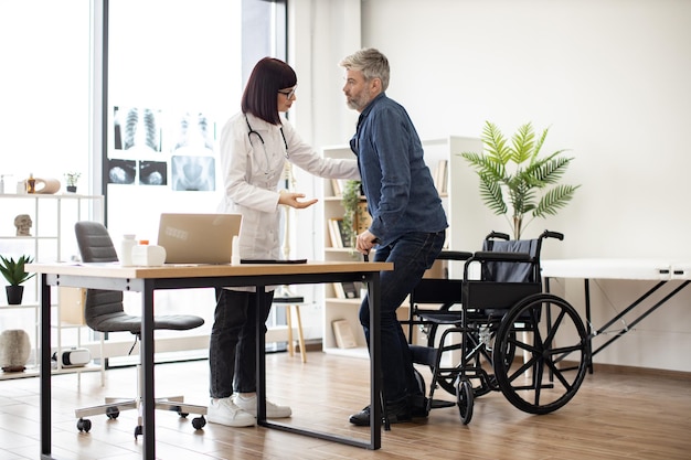 Médecin soutenant un homme tout en levant d'un fauteuil roulant à l'intérieur