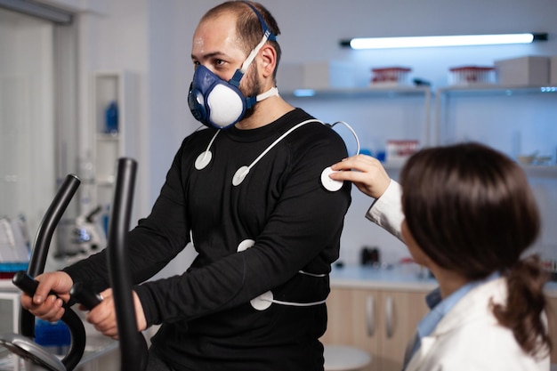 Médecin spécialiste mettant le capteur d'électrodes sur un sportif avec masque surveillant les données EGK dans un laboratoire d'examen médical. Homme athlète courant sur un entraîneur fintess travaillant à l'endurance du corps