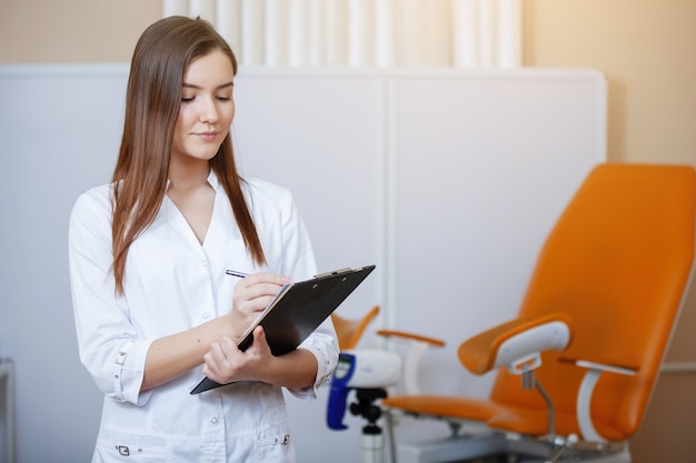 Médecin stagiaire jeune jolie femme en vêtements blancs avec une tablette dans ses mains posant à la caméra près de la chaise gynécologique orange