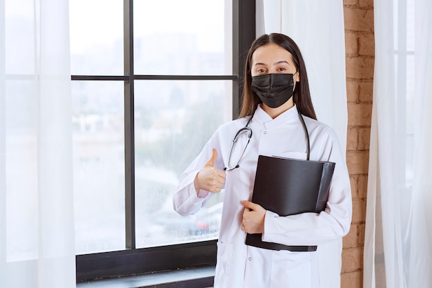 Médecin avec stéthoscope et masque noir debout à côté de la fenêtre et tenant un dossier d'histoire noire des patients et montrant un signe de plaisir.