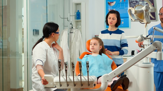 Médecin stomatologue expliquant à la mère le processus d'intervention dentaire pour les problèmes de dents d'un enfant, fille montrant à la masse affectée. Orthodontiste parlant à l'enfant s'asseyant sur la chaise stomatologique