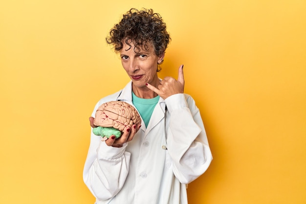 Médecin tenant un modèle de cerveau sur un studio jaune montrant un geste d'appel de téléphone portable avec les doigts