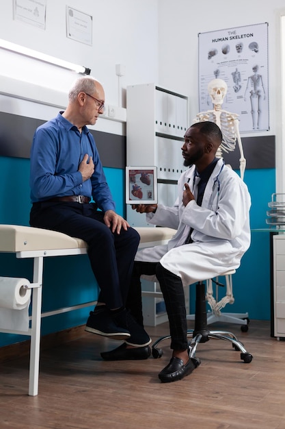 Médecin thérapeute afro-américain tenant une tablette avec radiographie cardiaque expliquant l'expertise en matière de maladie à un vieux patient à la retraite lors d'un rendez-vous clinique au bureau de l'hôpital. Notion de médecine