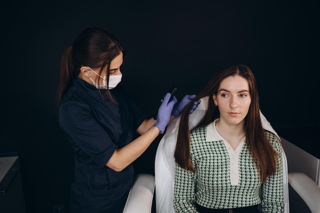 Un médecin trichologue examine les cheveux d'une patiente à l'aide d'un trichoscope en clinique Procédure de trichoscopie des cheveux Exécute un trichoscope sur le cuir chevelu et regarde le résultat sur l'écran du moniteur Trichoscopie informatique