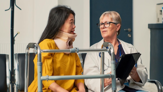 Médecin utilisant une tablette numérique pour consulter un patient avec une minerve cervicale, assis dans le hall de la zone d'attente. Assister à un rendez-vous médical avec un médecin, récupérer après un accident de fracture.