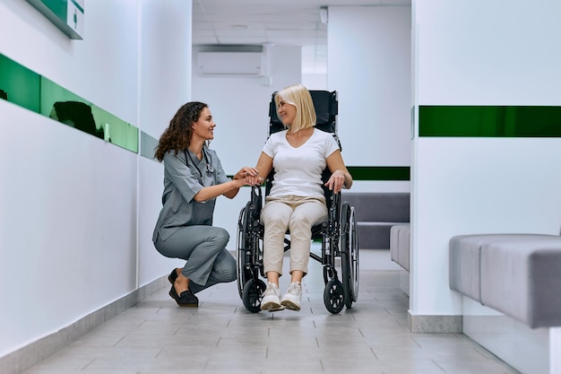 un médecin vêtu d'une combinaison médicale tient la main d'une femme dans le couloir de l'hôpital en fauteuil roulant et lui parle