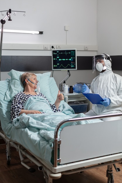 Médecin vêtu d'un costume ppe avec le visage shiled discutant avec un patient âgé, allongé au lit avec un masque à oxygène pendant l'épidémie de coronavirus