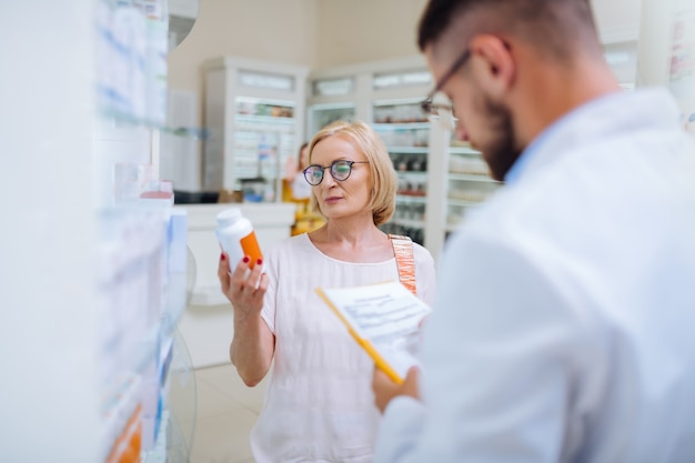 Médecine certifiée. Client mûr sérieux portant des lunettes tout en tenant une bouteille avec des vitamines