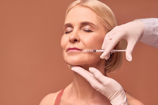 Photo médecine esthétiqueimage d'une femme adulte aux yeux fermés sur fond beige les mains dans des gants blancs et une blouse médicale font une correction de la forme de l'augmentation des lèvres par injection des lèvres