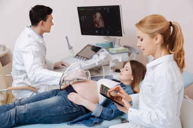 Des médecins qualifiés et qualifiés travaillant dans la clinique tout en effectuant un examen ultrasonique du ventre de femme enceinte et en utilisant des équipements et des gadgets modernes