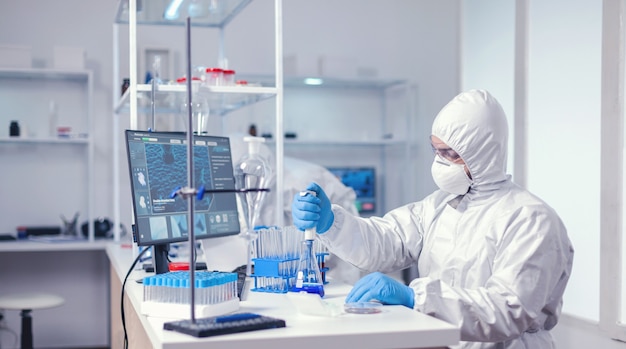 Medic portant un costume ppe dans une épidémie mondiale travaillant avec une micropipette dans un flacon en verre. Chimiste dans un laboratoire moderne faisant des recherches à l'aide d'un distributeur pendant l'épidémie mondiale de covid-19.