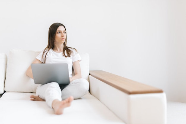 Méditant fille regardant loin assise sur un canapé moelleux avec un ordinateur portable blanc dans un salon lumineux. Une femme réfléchie tape du texte sur un ordinateur portable