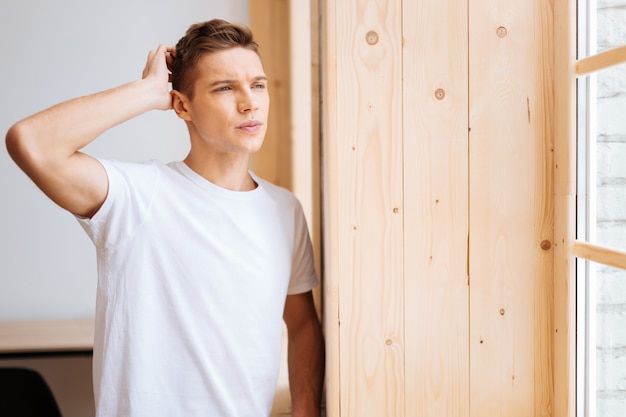 Méditer. Séduisant jeune homme bien construit inquiet pensant et touchant sa tête et regardant par la fenêtre