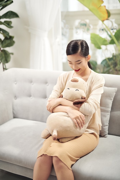 Meilleur ami à embrasser. Belle jeune femme asiatique étreignant son ours en peluche avec le sourire alors qu'il était assis sur un canapé
