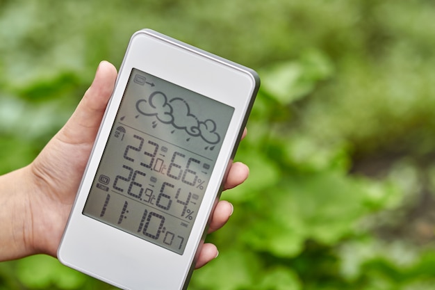 Meilleur appareil de station météo personnelle avec des conditions météorologiques à l'intérieur et à l'extérieur. Une fille tient un gadget à la main sur un fond de feuillage vert.