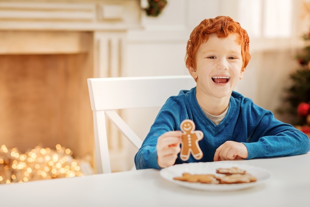 Le meilleur jour de ma vie. Heureux enfant rousse s'excitant et souriant largement tout en mangeant des hommes en pain d'épice à une table.