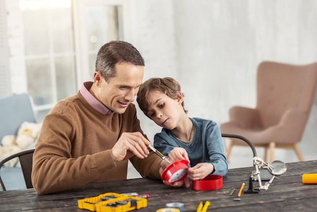 Meilleur père. Bel homme aux cheveux noirs heureux montrant des instruments à son fils assis sur la table et lui apprenant