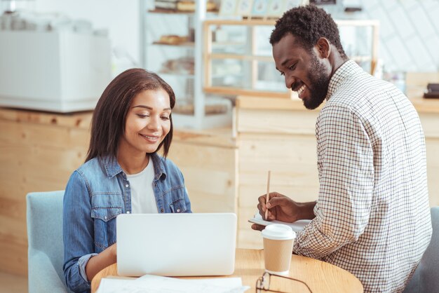 Meilleure coopération. Jolie jeune femme assise dans le café avec son collègue masculin et travaillant avec lui sur leur projet commun pendant que l'homme prend des notes