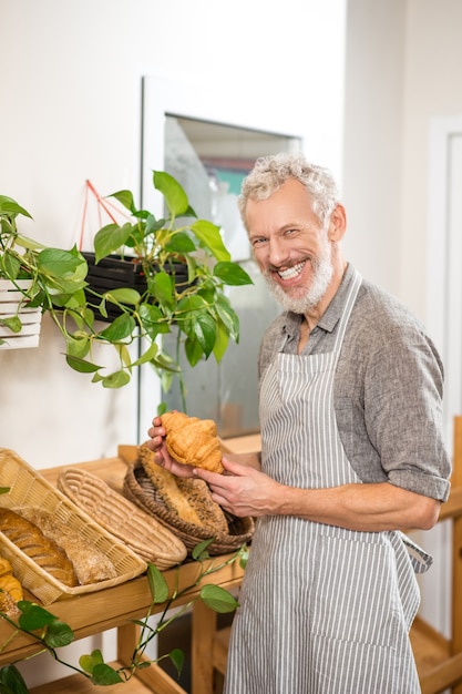 Meilleure cuisson. Ravissant homme aux cheveux gris barbu avec un croissant dans ses mains près de rack avec des pâtisseries en mini boulangerie