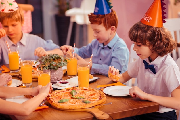 Meilleurs amis. Garçon aux cheveux bouclés sérieux assis à la table tout en mangeant de la pizza