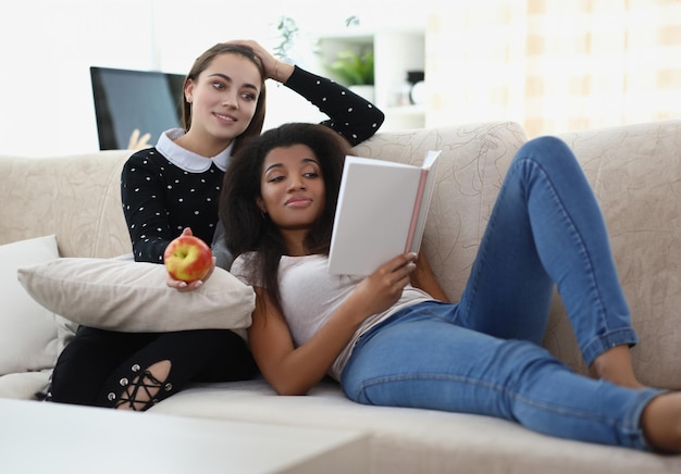 Photo meilleurs amis lisant un livre se reposant sur un canapé, pomme pour une collation, atmosphère relaxante