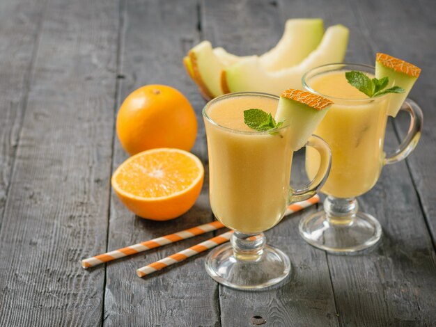 Melon, orange, tubes à cocktail et smoothies au melon dans des verres en verre sur une table rustique.