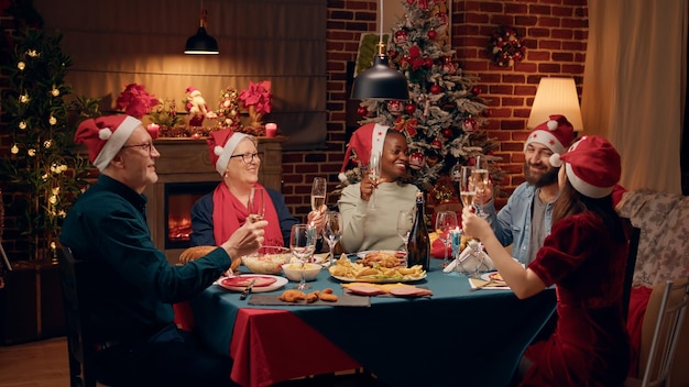 Membres de la famille heureux portant des chapeaux de fête tout en trinquant avec du champagne. Joyeuses personnes diverses riant de bon cœur tout en profitant du dîner de Noël ensemble à la maison.