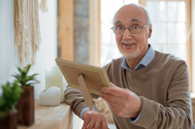 Une mémoire précieuse. Bel homme senior élégant portant cadre photo tout en portant des lunettes et regardant la caméra