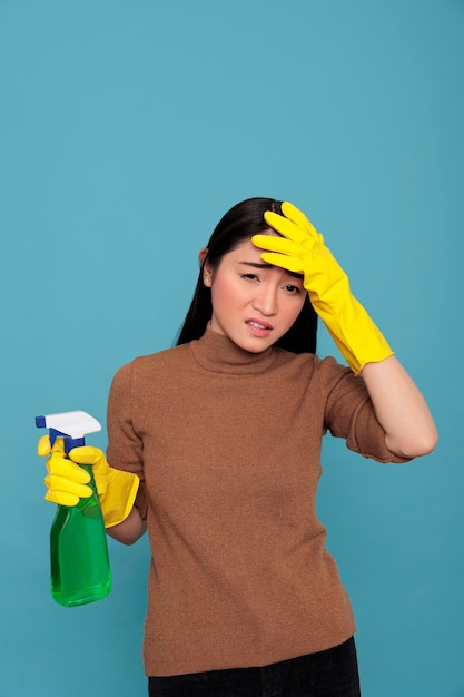Ménagère asiatique débordée de tâches ménagères quotidiennes tenant un pulvérisateur de détergent dans un gant en caoutchouc jaune isolé sur fond bleu, concept de maison de nettoyage, femme de ménage stressée frustrée