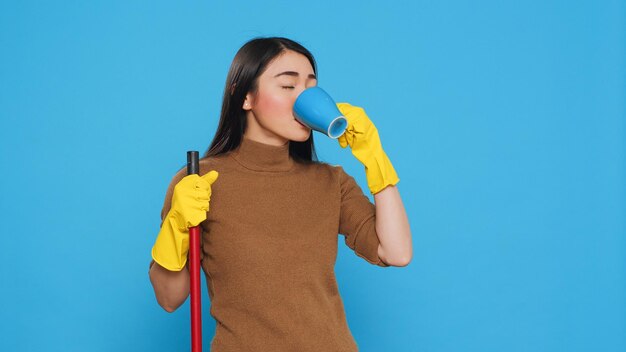 Ménagère joyeuse dégustant une tasse de café tout en nettoyant la maison avec un balai, debout en studio sur fond bleu. Femme de ménage utilisant un équipement de protection et des pratiques sanitaires pour assurer la sécurité des clients.