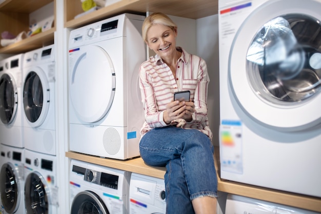 Ménagère moderne. Femme blonde en chemise rayée assis sur une machine à laver avec un smartphone à la main