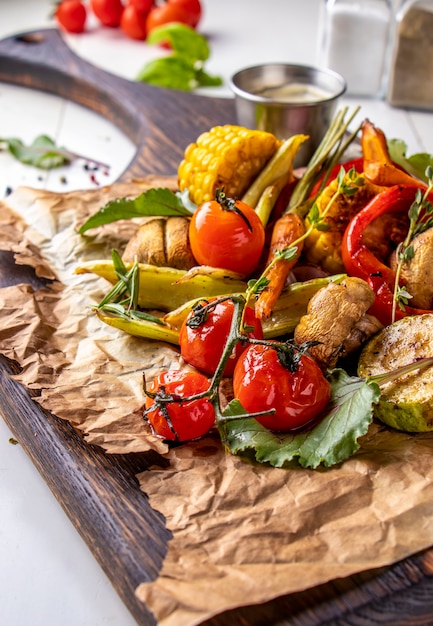 Menu Vegan Grill, Légumes grillés - courgettes, paprika, tomates cerises, maïs, carottes et champignons servis sur planche de bois à surface blanche, orientation verticale